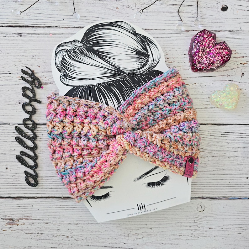 Puede incluir: Una diadema de crochet colorida en tonos rosa, azul y beige. La diadema est&aacute; anudada y se muestra sobre un fondo blanco con un dibujo de la cabeza de una mujer.