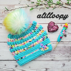 May include: A turquoise blue crochet hat with a rainbow and white pom pom. The hat has a striped pattern and is decorated with three heart-shaped buttons. The word "alliloopy" is written in black letters on a white background.