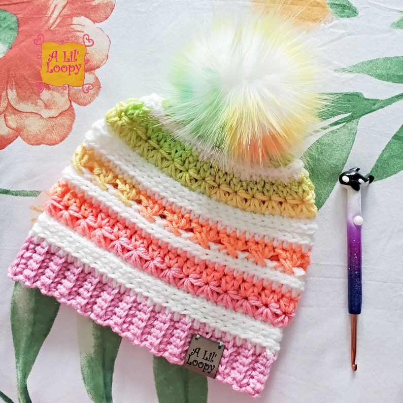 May include: A colourful crocheted hat with a white pom pom. The hat is striped with pink, white, orange, yellow and green. The hat has a label that says "A Lil' Loopy".