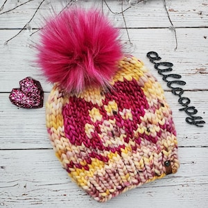 May include: A pink and yellow knitted beanie with a large pink faux fur pom pom. The beanie has a chunky knit texture and a leather patch on the side.