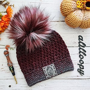 May include: A burgundy and gray ombre crochet beanie with a faux fur pom pom. The beanie has a leather patch with the text "In a world of ordinary, be extraordinary".