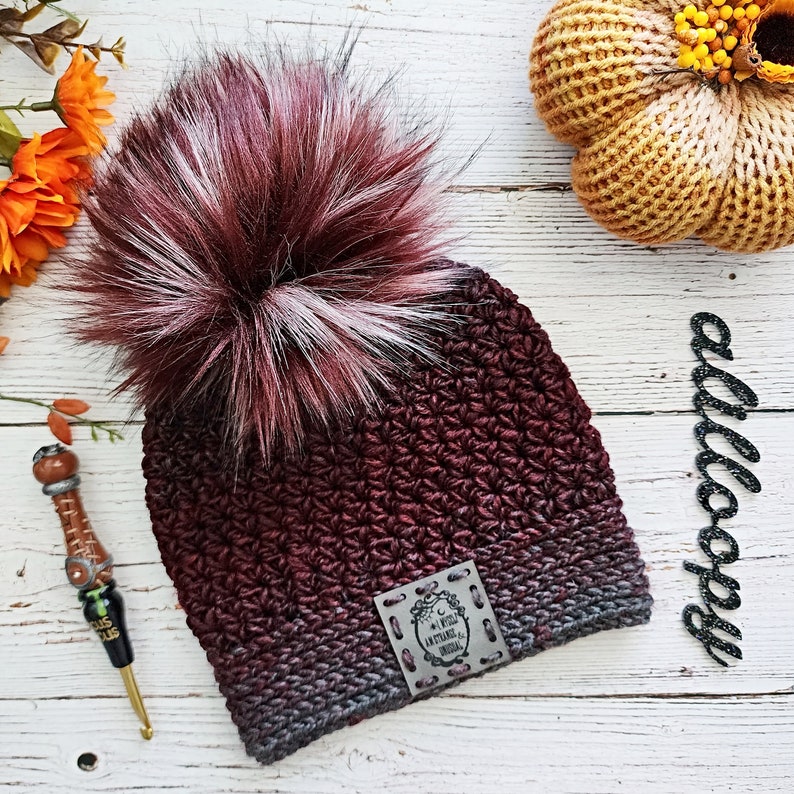 May include: A burgundy and gray ombre crochet beanie with a faux fur pom pom. The beanie has a leather patch with the text "In a world of ordinary, be extraordinary".