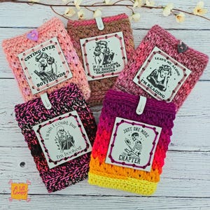 May include: Five colorful, crocheted book sleeves with attached tags. The sleeves are pink, brown, and purple, with text that reads "Crying Over Book Boyfriends", "If I'm Reading You Should Be Shhhhing", "Leave Me Alone I'm Reading", "I Wish I Could, But I'm Booked!", and "Just One More Chapter".