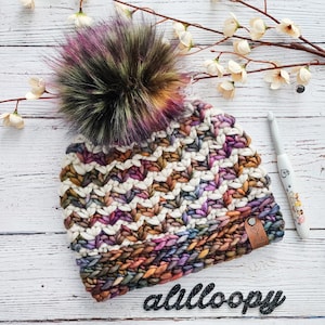 Op de afbeelding: Een kleurrijke gehaakte beanie met een meerkleurige nep bont pompon. De beanie heeft een chevron patroon en is gemaakt met een dik garen. De beanie heeft een bruin lederen label met de tekst "A Little Lovely" erop.