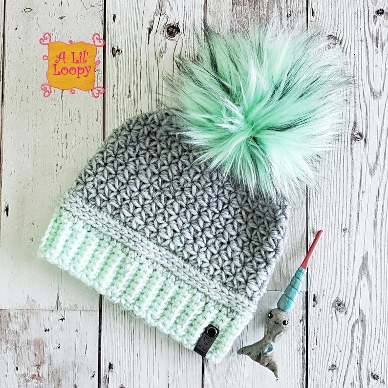 May include: A gray and mint green crochet beanie with a fluffy mint green pom pom. The beanie has a folded brim and a small black tag with the text "A Lil' Loopy" on it.
