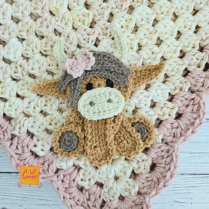 May include: A handmade crochet baby blanket featuring a Highland cow design. The blanket has a cream, pink, and tan color scheme. The cow has a light brown body, a gray hat with a pink bow, and a cream-colored muzzle. The blanket is made with soft yarn.