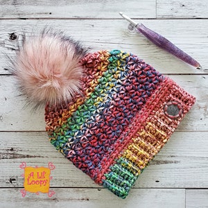 May include: A colourful crocheted beanie with a rainbow pattern and a fluffy pink pom pom. The beanie has a button on the side and is made with a variety of yarn colours.
