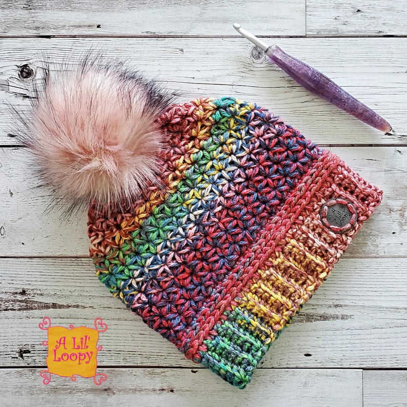 May include: A colorful crocheted beanie with a rainbow pattern and a fluffy pink pom pom. The beanie has a button on the side and is made with a variety of yarn colors.
