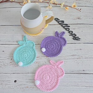 May include: Three crocheted bunny-shaped coasters in pink, purple, and teal with white pom-poms in the center. The coasters are on a white wooden surface.