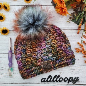 May include: A colorful crocheted beanie with a faux fur pom pom. The beanie has a brown leather patch with the text "Fall Babe" on it.