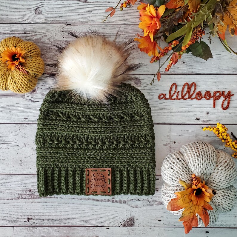 May include: A green crocheted beanie with a faux fur pom pom and a brown leather patch that says "Moons Out, Brooms Out". The beanie is on a white wooden surface with autumnal decorations.