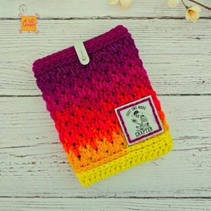 Pode incluir: Uma capa de livro de croch&ecirc; feita &agrave; m&atilde;o, num gradiente vibrante de cores, que vai do roxo escuro ao amarelo. Possui um fecho de press&atilde;o branco e uma etiqueta retangular com o texto "JUST ONE MORE CHAPTER" e um desenho de esqueleto.