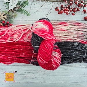 May include: A skein of thick, hand-dyed yarn featuring a gradient of vibrant red, soft pink, and charcoal gray tones. The yarn is twisted into a bundle, showcasing a variegated color pattern. A small orange tag with the text A Lil' Loopy is visible.