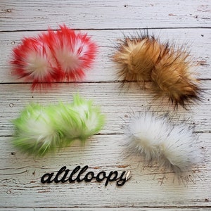 May include: Four fluffy faux fur pom poms in red, brown, green, and white with black tips. The pom poms are arranged on a white wooden surface. The word "atilloopy" is written in black cursive.