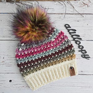 May include: A multi-colored crocheted beanie with a fluffy pom pom. The beanie has a cream colored brim and a brown leather tag with the text "A Little Loony" on it.