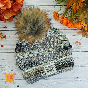 May include: A handmade crochet beanie in shades of gray, black, and tan, topped with a large faux fur pom-pom. The hat features a textured stitch pattern and a label that reads "Pumpkin Spice Girl."
