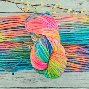 May include: A skein of colorful, chunky yarn in shades of pink, yellow, blue, and green. The yarn is arranged on a white wooden surface with a sprig of white flowers. The yarn is thick and appears soft.