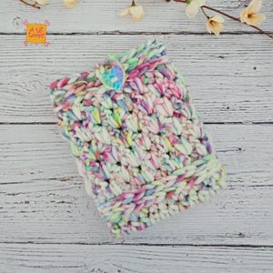 Pode incluir: Uma clutch multicolorida, tricotada &agrave; m&atilde;o, com fecho de bot&atilde;o. O fio &eacute; uma mistura de branco, rosa, amarelo e azul. A clutch &eacute; feita com um fio espesso e texturizado, e o bot&atilde;o tem formato de cora&ccedil;&atilde;o. O texto "A Lil Loopy" &eacute; vis&iacute;vel.