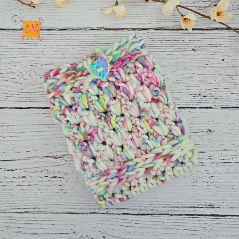 Pode incluir: Uma clutch multicolorida, tricotada &agrave; m&atilde;o, com fecho de bot&atilde;o. O fio &eacute; uma mistura de branco, rosa, amarelo e azul. A clutch &eacute; feita com um fio espesso e texturizado, e o bot&atilde;o tem formato de cora&ccedil;&atilde;o. O texto "A Lil Loopy" &eacute; vis&iacute;vel.