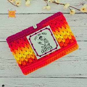 Pode incluir: Um aquecedor de pesco&ccedil;o de croch&ecirc; em tons vibrantes de rosa, laranja e amarelo. Uma etiqueta branca com o texto "JUST ONE MORE CHAPTER" e um esqueleto lendo um livro &eacute; fixada com um bot&atilde;o. O aquecedor de pesco&ccedil;o &eacute; feito &agrave; m&atilde;o.