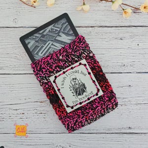 Pode incluir: Uma capa de leitor de e-books de croch&ecirc; rosa, preta e marrom com fecho de bot&atilde;o. A capa cont&eacute;m um leitor de e-books e apresenta uma etiqueta branca com um gr&aacute;fico de esqueleto e o texto "I wish I could, but I'm booked!"