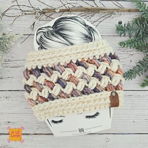 May include: A chunky knit headband in cream, lavender, peach, and brown tones. The headband features a woven design and a small leather tag. The product is displayed on a card with a drawing of hair.