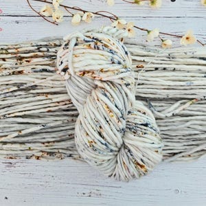 May include: A skein of thick, off-white yarn with brown and blue speckles, alongside a length of the same yarn. The yarn is arranged on a white wooden surface, with delicate white flowers in the background. The yarn is labeled "A Lil' Loopy".