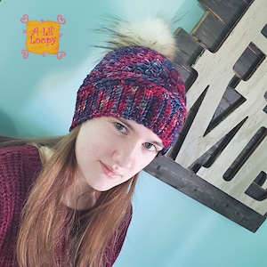 May include: A woman wearing a colorful knitted beanie with a faux fur pom pom. The beanie has a multi-colored pattern of red, blue, and purple.