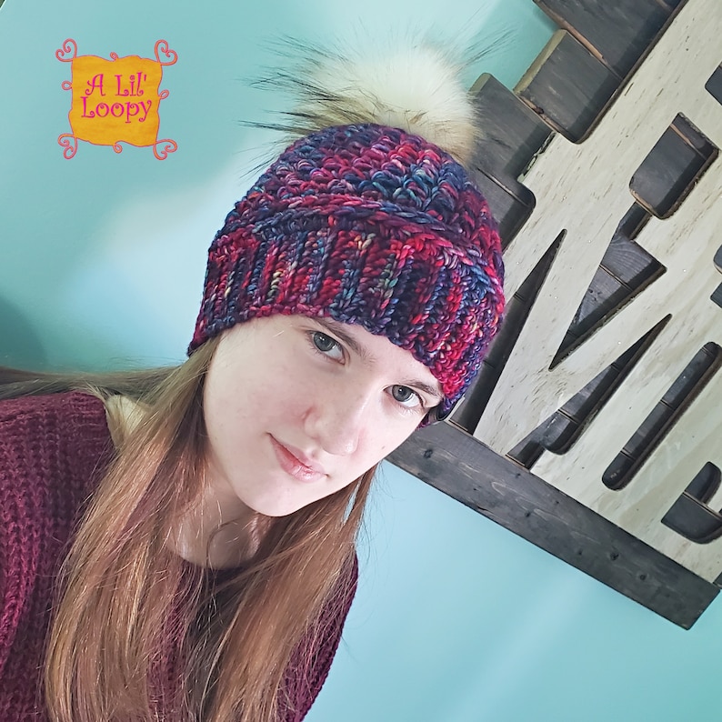 May include: A woman wearing a colorful knitted beanie with a faux fur pom pom. The beanie has a multi-colored pattern of red, blue, and purple.