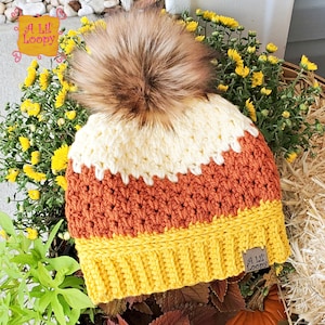 May include: A crocheted candy corn beanie hat with a brown faux fur pom pom. The hat is white, orange, and yellow. The hat has a brown leather tag with the text "A Lil' Loopy" on it.