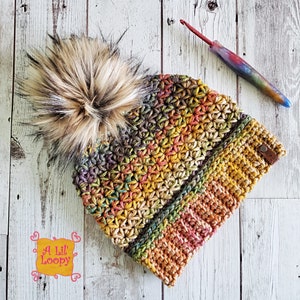 May include: A colorful crocheted beanie with a brown faux fur pom pom. The beanie has a multi-colored star stitch pattern and a brown leather tag with the text "A Lil' Loopy" on it.