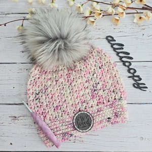 May include: A pink and white crocheted beanie with a faux fur pom pom. The beanie has a brown leather patch with the text "So Loved John 3:16".
