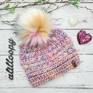 May include: A pink, purple and yellow crocheted beanie with a fluffy pom pom made of black, white and pink fur. The beanie has a brown leather tag with the word "alloopy" on it.