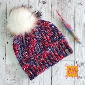 May include: A colorful crocheted beanie with a fluffy white pom pom. The beanie has a textured stitch pattern and a fold-over brim. The beanie is labeled "A Lil' Loopy".