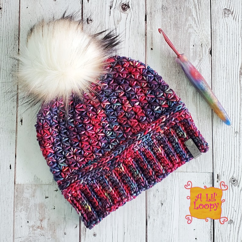 May include: A colorful crocheted beanie with a fluffy white pom pom. The beanie has a textured stitch pattern and a fold-over brim. The beanie is labeled "A Lil' Loopy".