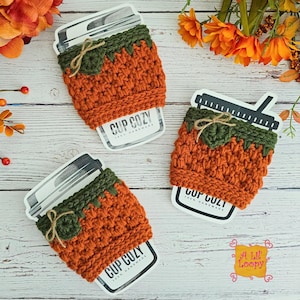 Peut inclure: Trois manchons à tasse en crochet orange et vert avec un motif de citrouille. Chaque manchon a un dessus vert et un petit nœud. Le texte "CUP COZY 100% HANDMADE" est visible sur la tasse. Fond en bois clair.