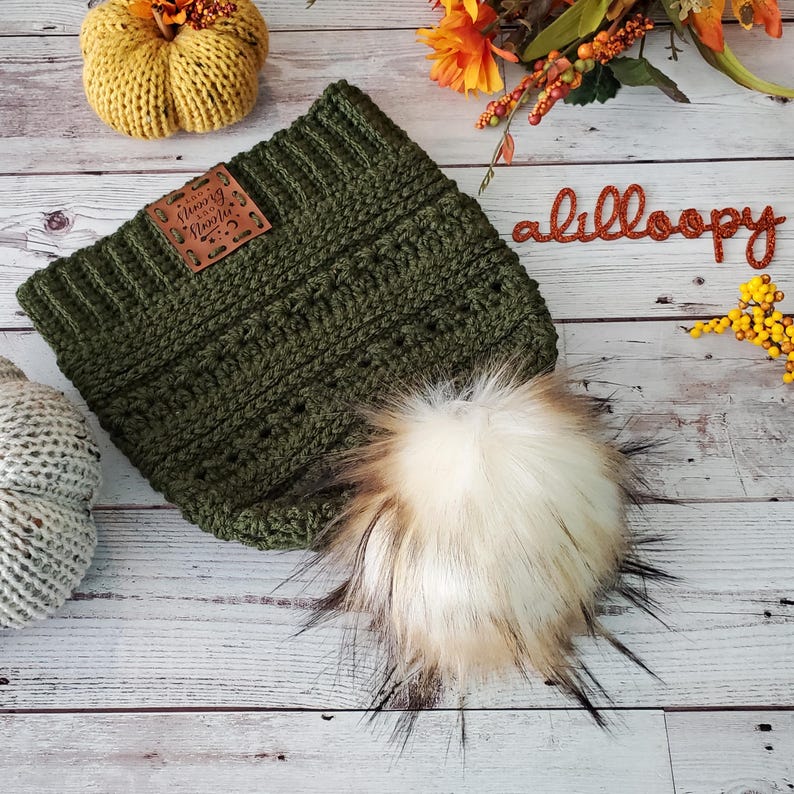 May include: A green crocheted beanie with a faux fur pom pom and a brown leather patch that says "Happy Place" on it. The beanie is sitting on a wooden surface with autumnal decorations.