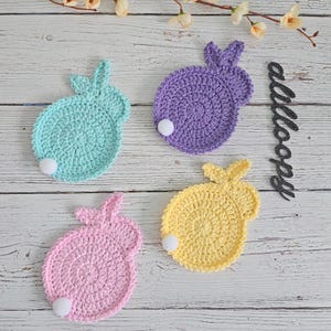 Może przedstawiać: Cztery dziergane podkładki pod kubki w kształcie królika w kolorze fioletowym, jasnoniebieskim, różowym i żółtym. Każda podkładka ma biały pompon w środku.