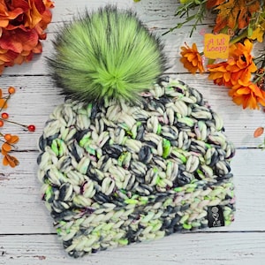 May include: A chunky knit beanie in cream, gray, and neon green with a large, fluffy green pom-pom. The hat features a textured stitch pattern and a small tag with the text "A Lil' Loopy".