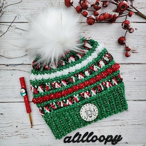Puede incluir: Un gorro de ganchillo con rayas horizontales de hilo verde, rojo y blanco con detalles en plata metálica. El gorro está rematado con un gran pompón de piel sintética blanca e incluye una etiqueta de madera redonda con el texto JOY y un reno.