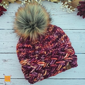 May include: A chunky knit beanie featuring a variegated yarn pattern in shades of deep purple, magenta, and bright orange. The hat has a thick braided brim and a large, fluffy faux fur pom-pom on top. A small brown leather tag is attached to the side.