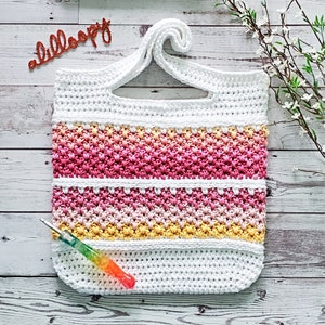 May include: White crochet tote bag with a pink, yellow, and white ombre stripe design. The bag has a single loop handle and a crochet hook is lying next to it.