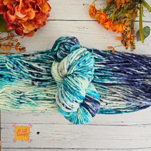 May include: A skein of chunky yarn in shades of blue, teal, and white, with dark blue speckles. The yarn is displayed on a white wooden surface, with orange flowers and foliage in the background. The yarn is labeled "A Lil' Loopy".