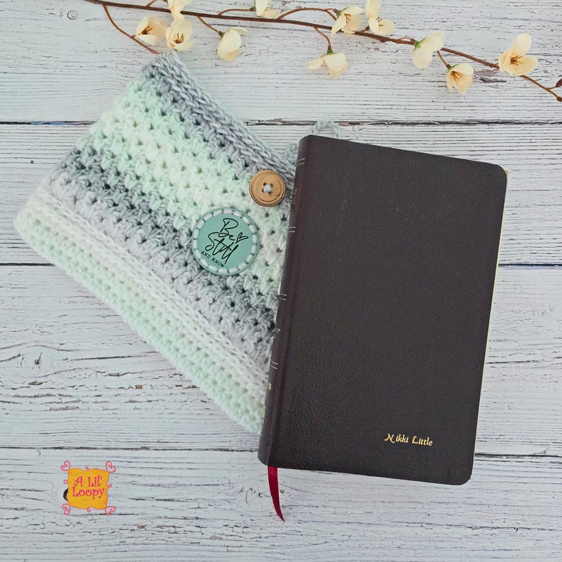 Pode incluir: Um livro de couro castanho com uma fita vermelha como marcador ao lado de uma capa de livro de croch&ecirc;. A capa &eacute; em tons de branco, cinza e verde menta, com um bot&atilde;o de madeira e um r&oacute;tulo turquesa que diz "Best Stash".