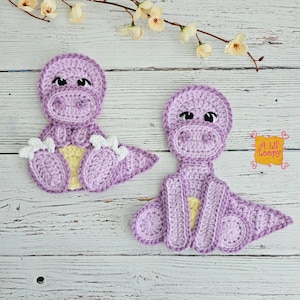 Pode incluir: Duas aplicações de dinossauros de crochê feitas à mão em tons de roxo, amarelo e branco. Os dinossauros têm detalhes, incluindo olhos, pés e barriga. As aplicações são colocadas em uma superfície de madeira branca com detalhes florais.