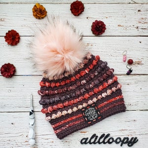 May include: A brown and pink crocheted beanie with a fluffy pom pom. The beanie has a black patch with the text "A Day Without Coffee Is Like... Just Don't Do It".
