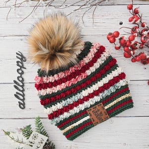 May include: A red, green, white, and cream striped crocheted beanie with a fluffy brown pom pom. The beanie has a brown leather patch with the text "Little Dog Tales" on it.