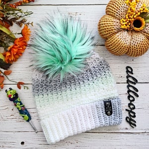 May include: A white and gray ombre crochet beanie with a light green faux fur pom pom. The beanie has a white ribbed brim and a small black leather tag with the text "A Little Lovely" on it.