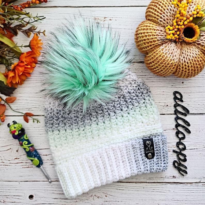 May include: A white and gray ombre crochet beanie with a light green faux fur pom pom. The beanie has a white ribbed brim and a small black leather tag with the text "A Little Lovely" on it.