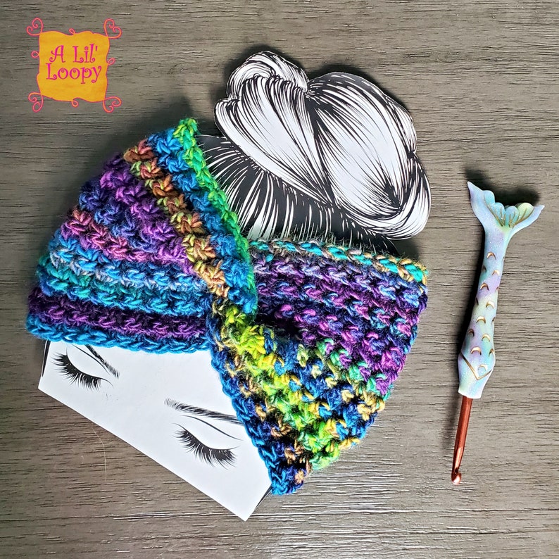 Puede incluir: Una diadema de crochet colorida en tonos azules, verdes y morados. La diadema est&aacute; doblada por la mitad y se encuentra sobre un fondo blanco con un dibujo de la cabeza de una mujer. El texto "A Lil' Loopy" es visible en la esquina superior izquierda de la imagen.